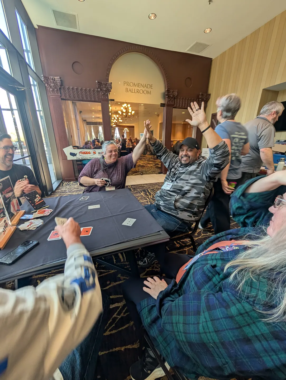 Two players high-five across a Sly3 game table while the others laugh