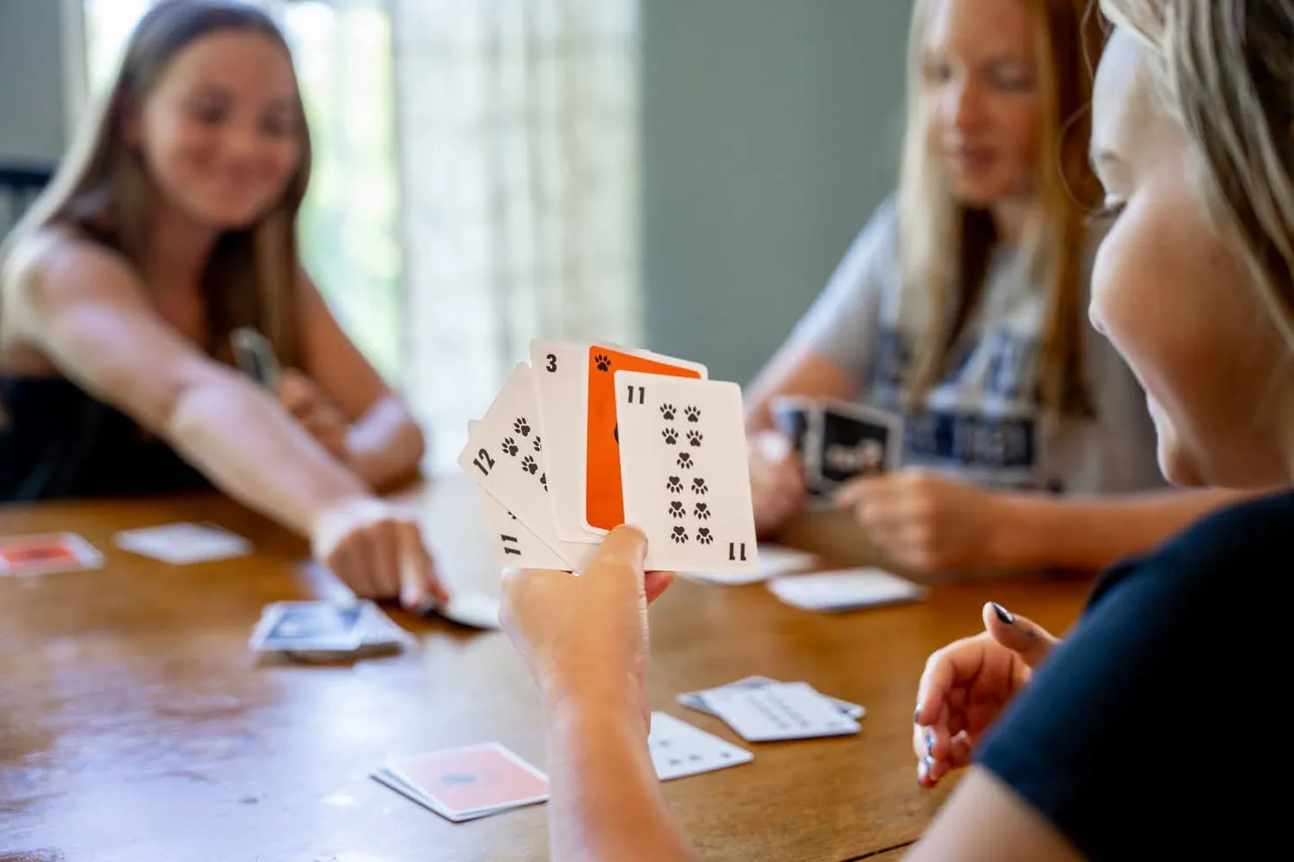 A hand holds a fan of Sly3 cards toward the camera with other players behind
