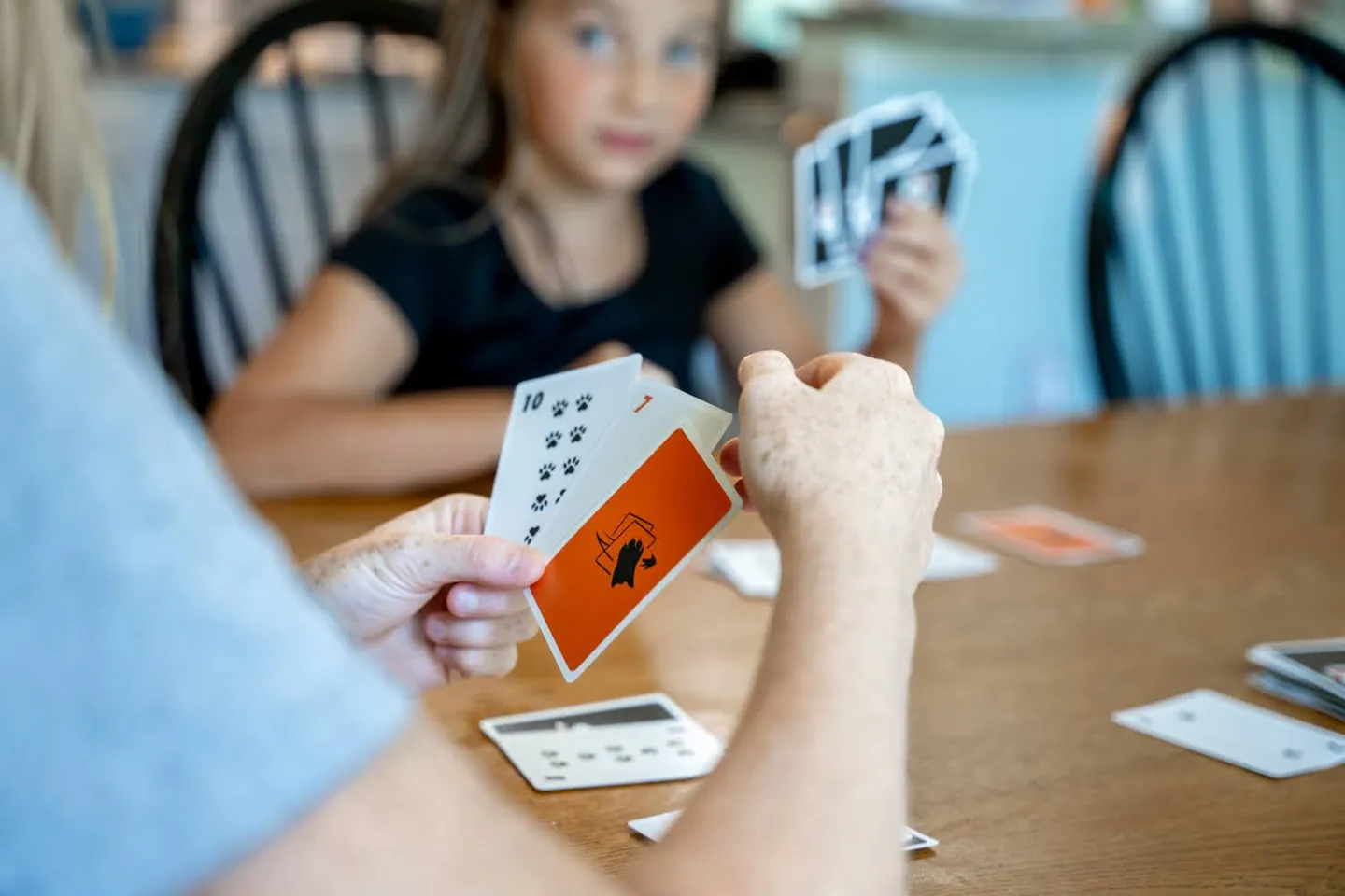 An adult holds a fan of Sly3 cards while a child across the kitchen table holds hers