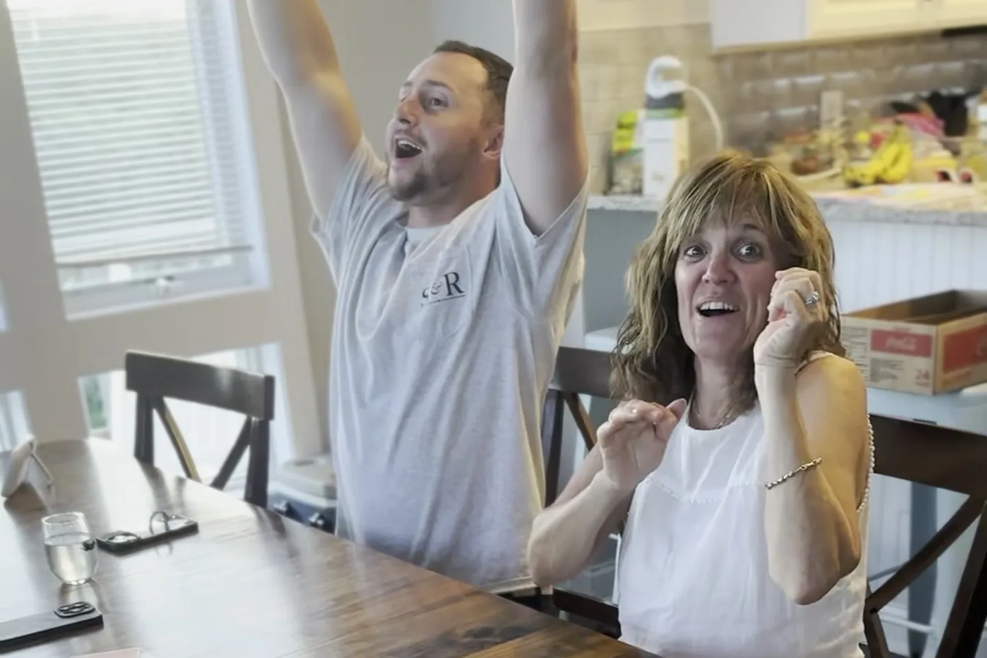 A grandmother laughing with her fist up at the Sly3 table while her son celebrates beside her, mid-round