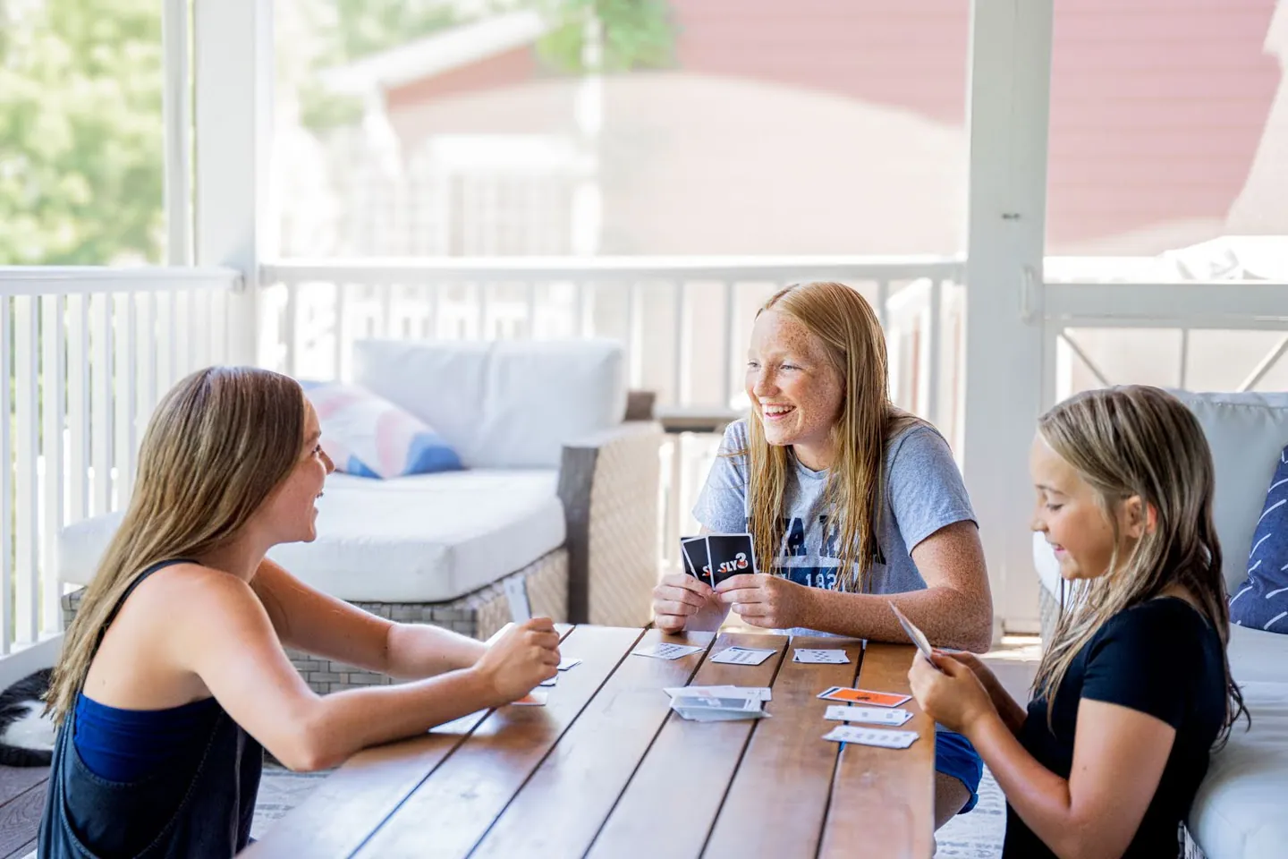 Three young players mid-game around a porch table, one holding up her Sly3 cards